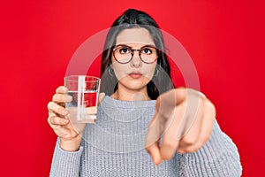 Young beautiful woman drinking a glass of fresh water over isolated red background pointing with finger to the camera and to you,