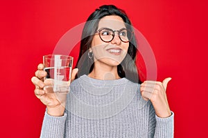 Young beautiful woman drinking a glass of fresh water over isolated red background pointing and showing with thumb up to the side