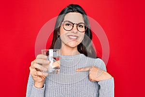 Young beautiful woman drinking a glass of fresh water over isolated red background with surprise face pointing finger to himself