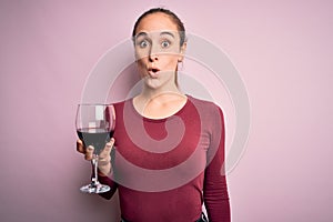 Young beautiful woman drinking glass with red wine over isolated pink background scared in shock with a surprise face, afraid and