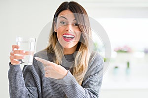 Young beautiful woman drinking glass of water at home very happy pointing with hand and finger