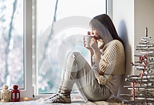 Young beautiful woman drinking hot coffee sitting on window sill photo