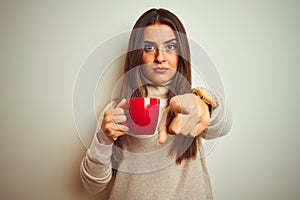 Young beautiful woman drinking red cup of coffee standing over isolated white background pointing with finger to the camera and to