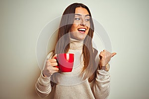 Young beautiful woman drinking red cup of coffee standing over isolated white background pointing and showing with thumb up to the