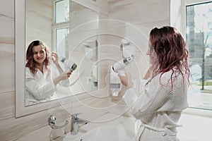 Young beautiful woman with dyed hair in white bathrobe in bathroom dries hair with hairdryer and smiles.
