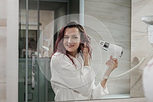 Young beautiful woman with dyed hair in white bathrobe in bathroom dries hair with hairdryer and smiles.