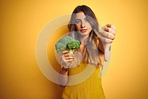 Young beautiful woman eating broccoli over yellow  background with angry face, negative sign showing dislike with thumbs