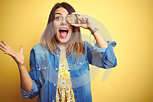 Young beautiful woman eating chocolate chips cookie over yellow background very happy and excited, winner expression celebrating