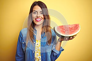 Young beautiful woman eating fresh healthy watermelon slice over yellow background with a happy face standing and smiling with a