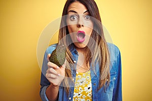 Young beautiful woman eating healthy avocado over yellow background scared in shock with a surprise face, afraid and excited with