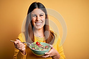 Young beautiful woman eating healthy fresh salad over yellow background with a happy face standing and smiling with a confident