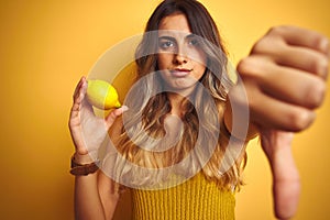Young beautiful woman eating a lemon over yellow isolated background with angry face, negative sign showing dislike with thumbs