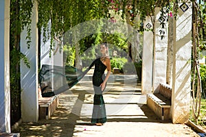 Young beautiful woman in elegant dress poses for photo among vines hanging from the columns in the park. The woman is happy and