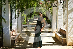 Young beautiful woman in elegant dress poses for photo among vines hanging from the columns in the park. The woman is happy and