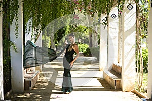 Young beautiful woman in elegant dress poses for photo among vines hanging from the columns in the park. The woman is happy and