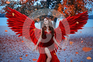 Young beautiful woman fallen angel stands in the park near a tree with orange falling leaves. Creative red suit, huge