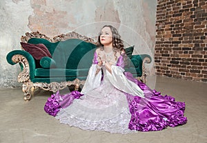 Young beautiful woman in fantasy white and purple rococo style medieval dress sitting on the floor and praying