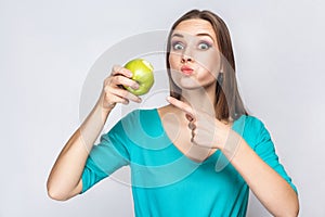 Young beautiful woman with freckles and green dress holding and eating apple and pointing and looking at camera with big eyes.