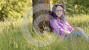 Young beautiful woman in glasses with wireless white headphones sitting on green grass near tree in summertime. Pretty