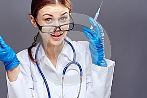 Young beautiful woman on a gray background in a medical dressing gown and gloves holds a syringe, medicine, doctor