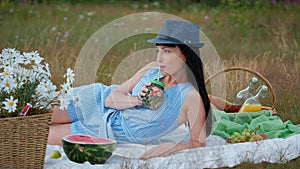 A young beautiful woman in a hat and dress is drinking lemonade from a can while sitting on a plaid on the green grass