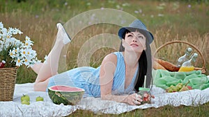 A young beautiful woman in a hat and dress is drinking lemonade from a can while sitting on a plaid on the green grass