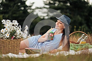 A young beautiful woman in hat and dress is drinking lemonade from a glass jar, sitting on plaid on green grass. Picnic