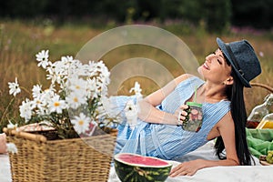 A young beautiful woman in hat and dress is drinking lemonade from a glass jar, sitting on plaid on green grass. Picnic