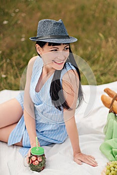 A young beautiful woman in hat and dress is drinking lemonade from a glass jar, sitting on plaid on green grass. Picnic