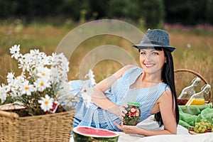 A young beautiful woman in hat and dress is drinking lemonade from a glass jar, sitting on plaid on green grass. Picnic