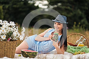 A young beautiful woman in hat and dress is drinking lemonade from a glass jar, sitting on plaid on green grass. Picnic