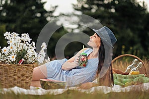 A young beautiful woman in hat and dress is drinking lemonade from a glass jar, sitting on plaid on green grass. Picnic