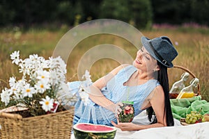 A young beautiful woman in hat and dress is drinking lemonade from a glass jar, sitting on plaid on green grass. Picnic