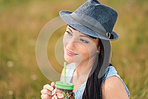 A young beautiful woman in hat and dress is drinking lemonade from a glass jar, sitting on plaid on green grass. Picnic