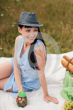 A young beautiful woman in hat and dress is drinking lemonade from a glass jar, sitting on plaid on green grass. Picnic