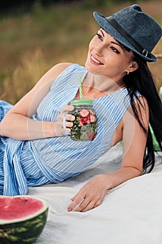 A young beautiful woman in hat and dress is drinking lemonade from a glass jar, sitting on plaid on green grass. Picnic