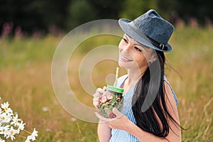 A young beautiful woman in hat and dress is drinking lemonade from a glass jar, sitting on plaid on green grass. Picnic