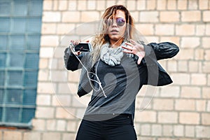 Young beautiful woman with headphones and smartphone in hand in mirrored sunglasses, a black leather jacket, black jeans danc