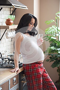 A young beautiful woman in her checkered pajamas is drinking a glass of pure water in the kitchen. Beautiful healthy pregnancy and