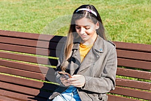 Young beautiful woman hipster cheerfully smiling and texting via smartphone sitting on bench in park in spring