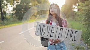 Young beautiful woman hitchhiking standing on the road holding adventure sign. Summer time.