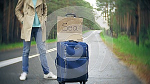 Young beautiful woman hitchhiking standing on the road with a suitcase on a table with an inscription SEA