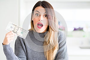 Young beautiful woman holding 20 dollars bank note at home scared in shock with a surprise face, afraid and excited with fear