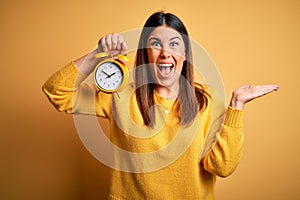 Young beautiful woman holding alarm clock standing over isolated yellow background very happy and excited, winner expression