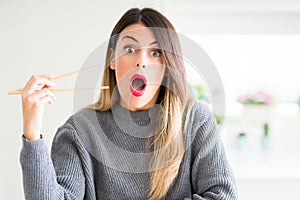 Young beautiful woman holding asian chopsticks at home scared in shock with a surprise face, afraid and excited with fear