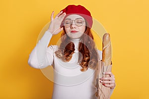 Young beautiful woman holding bag of fresh baguette over yellow background, poses with hand on head for mistake, remember error,