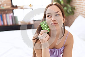 Young beautiful woman holding birth control pills sitting on bed at bedroom