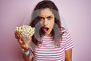 Young beautiful woman holding bowl with pistachios over isolated pink background scared in shock with a surprise face, afraid and