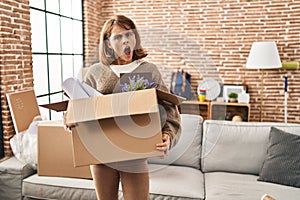 Young beautiful woman holding box moving to a new home in shock face, looking skeptical and sarcastic, surprised with open mouth