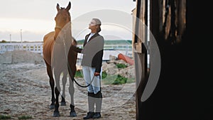 a young beautiful woman holding a brown horse by a leash in front of a stable